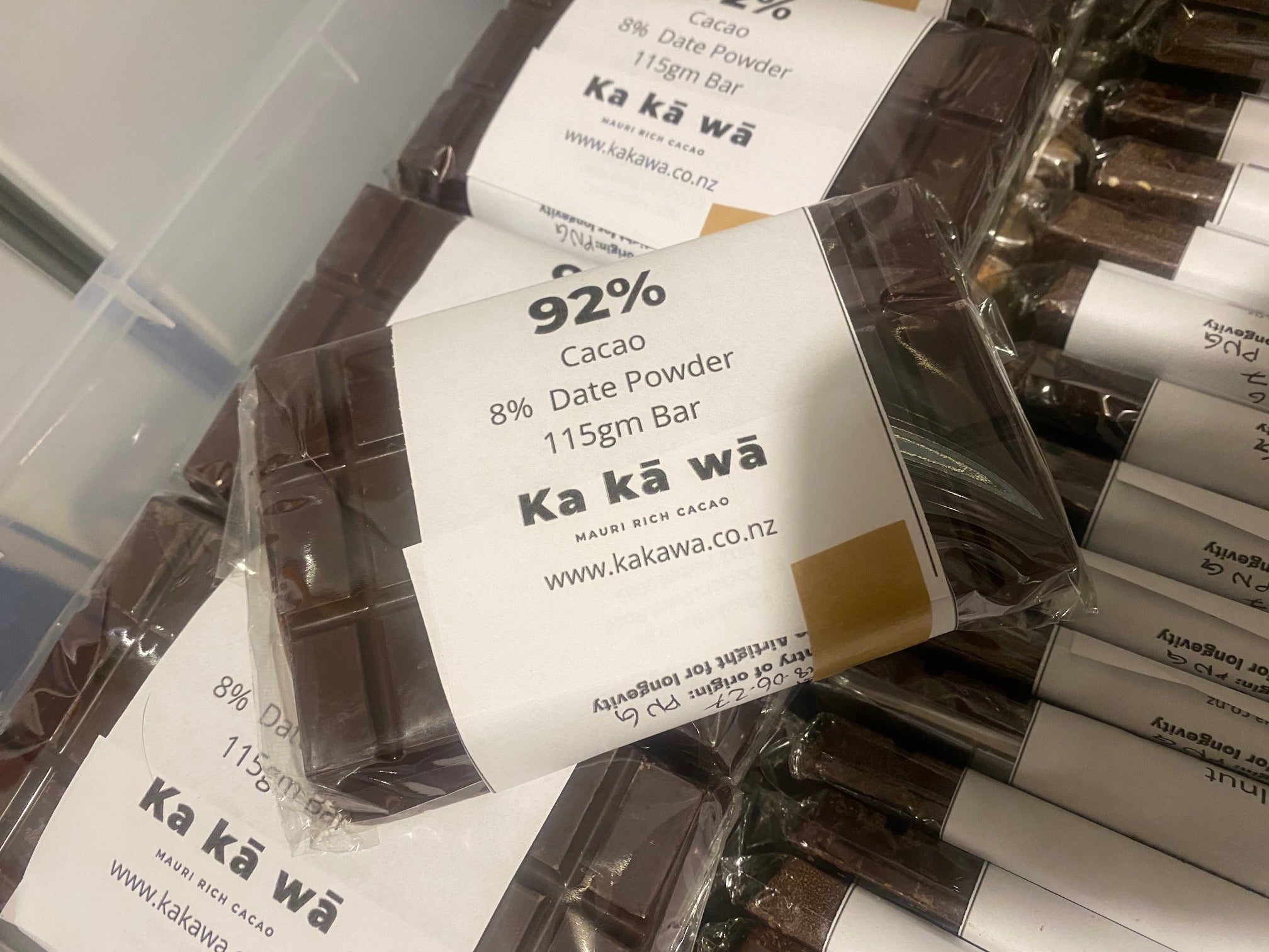 Several packaged chocolate powder bars stacked together. The front package is clear plastic with a white label reading 'Ka kā wā' and '92% cocoa 8% cocoa powder 115g bar'.